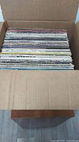 View into a cardboard box showing a large collection of vinyl LP records stacked vertically, album covers visible from the side varying in color and condition.