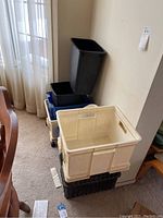 Photo showing stacked bins and one tall black garbage can near a white wall and window curtain.