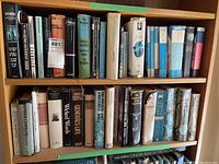 Books on wooden shelves showing multiple hardcover and paperback books including boxed sets and loose volumes. Spines visible with some wear and protective plastic coverings on some books. Titles include classic literature, mystery, and fantasy series.
