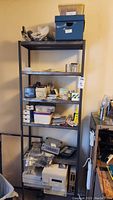 Black metal shelving rack filled with various aged computer hardware parts, components, boxes, and accessories in assorted condition.