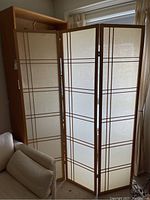 Three-panel room divider unfolded partly, showing wooden frame and beige fabric with dark brown lines.