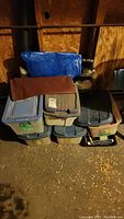 Plastic storage bins stacked against wall with painting supplies inside and some covers placed on top.