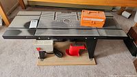 Photo of router table showing metal surface with grooves, adjustable fences, power switch with red button mounted on base, wooden platform base under table.