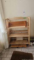 Wooden shelf unit with three shelves, vertical sides, decorative top, placed against wall indoors. Signs of dirt and wear visible on shelves and floor below.