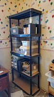 Full view of black plastic shelving unit with four tiers, standing in a room with balloon-patterned wallpaper.