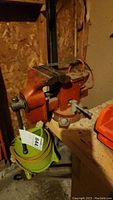 Orange metal workbench vise mounted on wooden bench, showing side profile and screw handle.