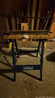 Front view of the work bench showing wooden top with holes and yellow clamps, blue frame, and metal pipes on top.
