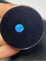 Close-up of a round opal gemstone showing its vibrant blue and green colors on black foam inside a plastic container, held in hand.