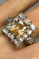 Close-up front view of sterling silver ring with yellow rectangular center stone and clear crystals surrounding it on finger.