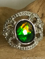 Close-up of sterling silver ring with oval Ammolite gemstone showing vibrant green and orange colors, ornate silver setting with small stones or detailing.