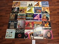 Top-down image of 25 vintage LP vinyl records laid out on wooden floor, showcasing diverse album covers including orchestra music, jazz, soundtracks, and pop artists.