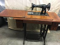 Full view of the antique Singer sewing machine attached to the wooden foldable table with drawers and cast iron treadle base.