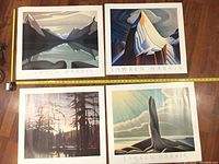 Four unframed lithograph prints by Lawren Harris arranged on wooden floor with measuring tape, showing scenic landscapes including mountains and forest.
