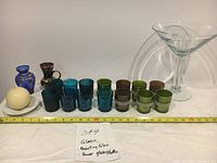 Lot arranged with ruler showing size, includes clear martini glass, various colored small glasses, two decorative bottles, a spherical candle, and a plate.