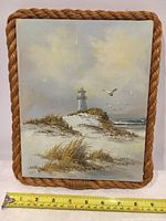 Frontal view of rope-framed oil painting showing coastal scene with lighthouse and seagulls. Tape measure for size reference placed in front.