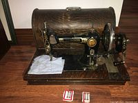Front view of the black cast iron hand-crank sewing machine on wooden base with wooden carry case behind it. Machine is threaded and has some fabric under the needle. Two packages of spare needles or threads are placed in front.