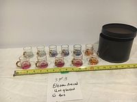 Eleven pressed shot glasses in assorted colors with gold handles displayed next to a black cylindrical carrying case, a tape measure for size reference, and a handwritten label noting eleven pressed shot glasses with case.