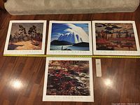 Four lithographs laid out on a wooden floor showing artwork and artist names: Tom Thomson, Lawren Harris, J.E.H. MacDonald, and A.Y. Jackson.