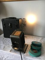 Full view of vintage Kodak KODASCOPE EIGHT model 70 film projector, black textured carrying case behind, and three rolls of 8mm film in teal green metal canisters on table.