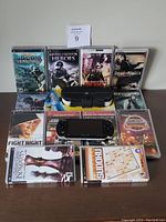 Photo showing Sony PSP console, black carrying case, batteries, and 14 game cases arranged on a wooden surface.
