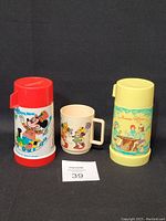 Photo of two vintage thermal cups by Aladdin Industries and one Disney plastic cup by Deca on a black background, showing the colorful character and theme designs.