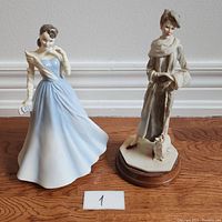 Two Royal Doulton porcelain figurines displayed on wood floor against wall, one in blue dress, the other in winter attire with dog on base.