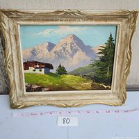 Frontal photo showing the entire framed painting with mountain landscape and cabin, including part of measuring tape to indicate size