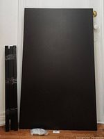 Black desk tabletop laying flat against a wall with a small bag of hardware in front and four black legs taped together to the side.