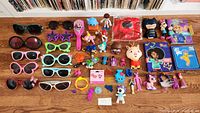 Photo showing a collection of assorted kids toys including action figures, dolls, and small character toys alongside a variety of children's sunglasses in different styles and colors on a wooden floor.