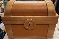 Closed tan plastic toy chest with rounded lid and keyhole detail on the front.
