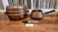 Set of copper cookware including pot with handle, pan, and mold on wood floor with marker for scale