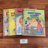 Four vintage Little Golden Books including Donald Duck, Winnie-the-Pooh, Mickey Mouse and Goofy, and Dumbo, arranged fanned out on wooden floor.