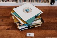 Stack of over 40 vintage 7-inch vinyl records in assorted colored paper sleeves on wooden floor with lot number sign (41).