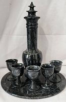 Front angled view of black etched floral marble decanter with lid, six pedestal goblets, and round tray on white backdrop.