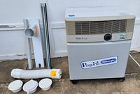 Front and side view of Delonghi Pinguino portable air conditioner with accessories laid out including exhaust hose, vent covers, and window vent parts.