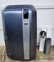Front view of Delonghi portable air conditioner unit with vent hose and adapter accessories on ground.