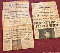 Stack of newspapers on a red fabric surface, showing The Washington Post and The Evening Star with prominent JFK assassination headlines.