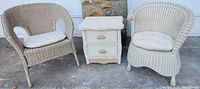Shows two white wicker curved back chairs with tufted cushions flanking a white wicker two-drawer dresser with shell-shaped wooden drawer pulls and scalloped detail on the top drawer front. Setting outside with stone wall background.