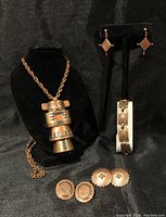 Full view of the lot showing the copper totem necklace on a chain, three pairs of copper earrings (two pairs of round studs and one pair of drop earrings), and a copper bracelet on display stands against a black background.