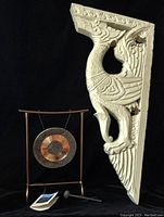 Photo showing the carved wooden corbel with a dragon/rooster/duck motif beside the gong and stand with mallet below, against a black background.