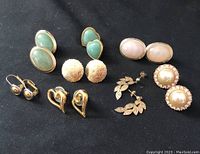 Top view of all nine pairs of earrings laid out on black fabric, showing variety of styles and materials including stone and gold-tone metal.