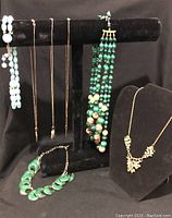 Seven vintage necklaces displayed on black necklace stand and platform, showing various styles including turquoise beaded, green enamel, and gold-tone chains.