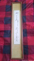 Frontal view of the antique Asian calligraphy painting in the wooden frame laid on a red and black checkered fabric surface.