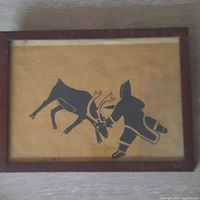 Frontal view of vintage Inuit print on jute showing dark silhouette of hunter and caribou in wooden frame.