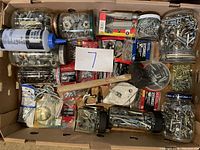 Top-down view of a cardboard box filled with assorted hardware items including screws, bolts, a small mallet, chalk line and chalk powder.