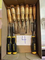 Top-down view of 10 chisels in cardboard box showing three DeWalt brand chisels and seven wooden handle chisels with varied blade sizes and some rust.