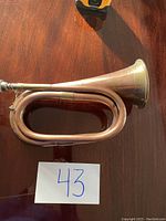 Copper and brass bugle lying on a wooden surface with a label '43'. Shows side view highlighting the coiled shape and bell.