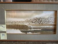 Full view of framed art showing large flock of geese flying over lagoon in brown tones, with fabric mat and gold inner border.