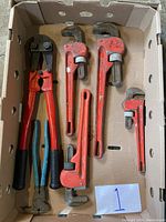 Box containing five red heavy duty pipe wrenches of various sizes and two tools with handles, one blue, one red, showing good used condition.