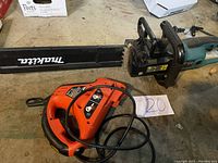Makita UC-4030A electric chain saw and Black & Decker jigsaw shown side by side on ground.